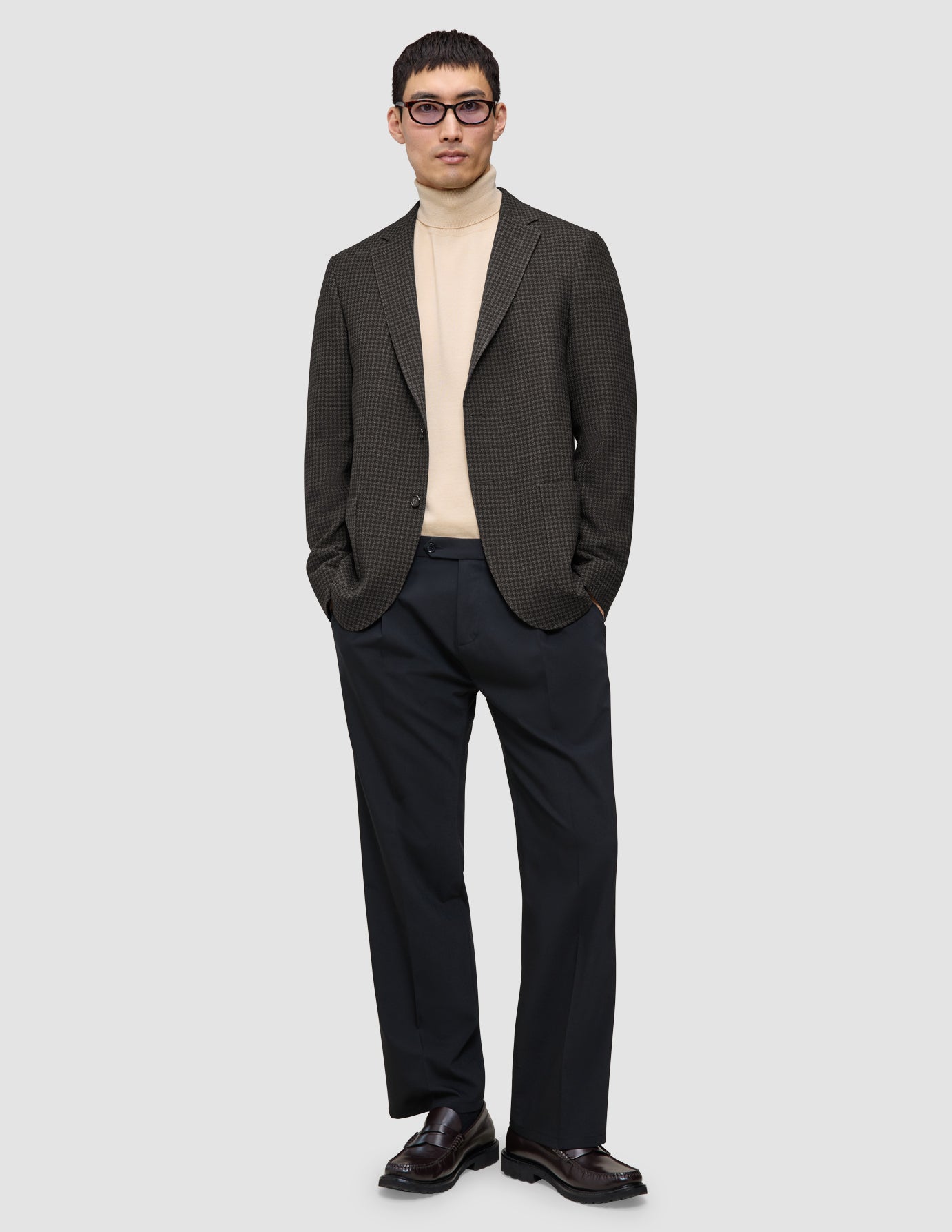 En mand har en mørkebrun, slim-fit Timeless-uldsblazer med tern på, over en beige rullekravebluse og mørke bukser.